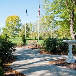 A scenic garden pathway featuring a bench, birdbath, and flags amidst lush greenery.