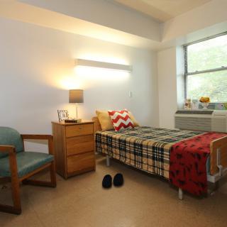 A cozy dorm room with a single bed, chair, and small dresser beside a window.