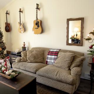 A cozy living room featuring a sofa adorned with a holiday-themed pillow, guitars mounted on the wall, and a decorated Christmas tree adding festive charm.