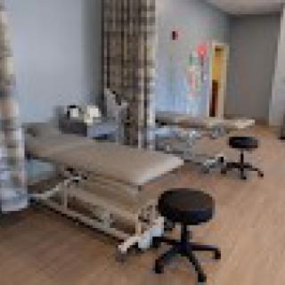 A healthcare examination room with multiple examination tables and stools, separated by curtains.