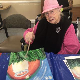 An elderly woman wearing a pink hat is painting a picture at a table.