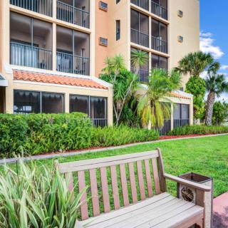 A peaceful view of an apartment building with a bench surrounded by lush greenery and palm trees.