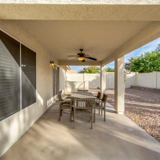 A peaceful covered patio with a dining table and ceiling fan, overlooking a sunny backyard.
