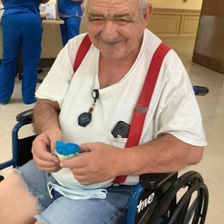 An elderly man in a wheelchair, smiling and holding a small container.