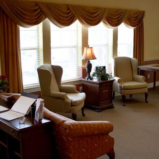 A cozy living room with armchairs, a table, and a lamp near large windows with draped curtains.