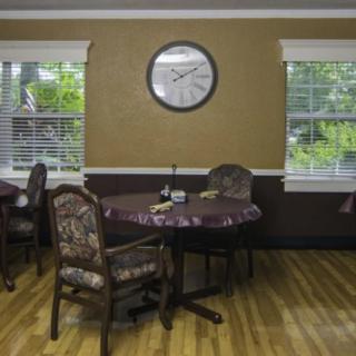 A cozy dining room with tables and chairs, featuring a clock on the wall and windows overlooking greenery.
