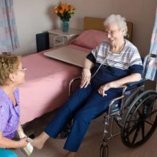 A caregiver attends to a senior woman in a wheelchair in a nursing home room.
