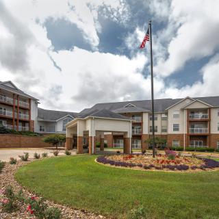 A multi-story apartment building with a landscaped garden and flagpole at the entrance under a cloudy sky.