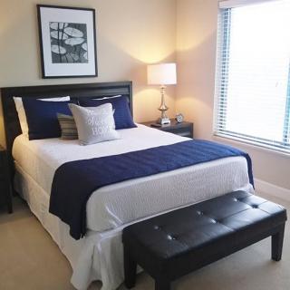 A modern bedroom with a neatly made bed, featuring blue and white bedding, a bedside lamp, and a cushioned bench at the foot of the bed.