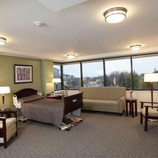 A well-lit hospital room with large windows, featuring a bed, seating area, and modern furnishings.