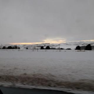 A serene winter landscape with a blanket of snow under a cloudy sky as the sun sets in the distance.
