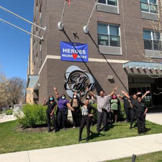 A group of people celebrating outside a building with flags and a banner that says 'HEROES WORK HERE'.