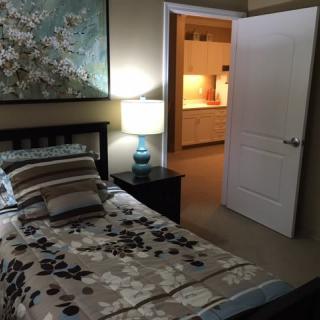A cozy bedroom with a bed, nightstand, and lamp, leading to an open door with a view of the adjacent room.