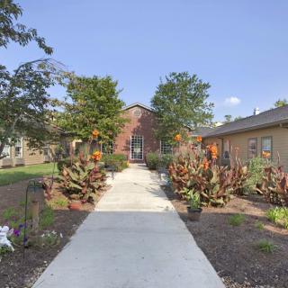 A landscaped pathway leads to a brick building, surrounded by trees and bushes under a clear sky.