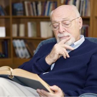 An elderly man wearing glasses is deep in thought while reading a book in a library.