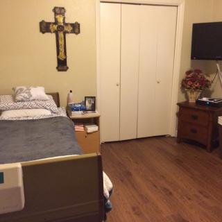 A small bedroom with a hospital bed, crucifix on the wall, TV, and a wooden side table.