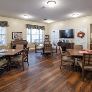 A communal dining area with wooden flooring, tables, and chairs, featuring a television on the wall.