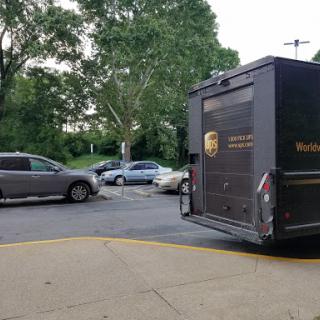 A UPS delivery truck parked in an urban setting with multiple cars and greenery in the background.