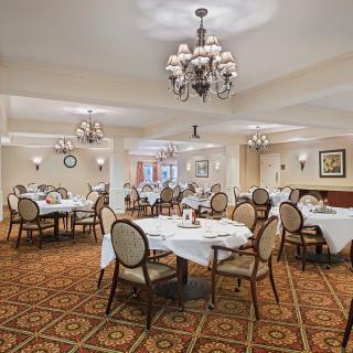 An elegant dining room with chandeliers, featuring several empty tables with white tablecloths and a patterned carpeted floor.