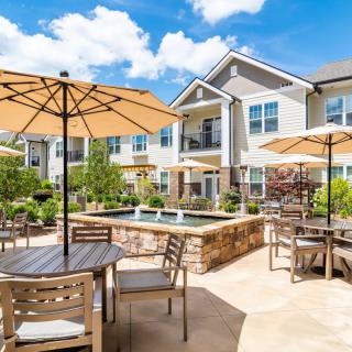 A sunny outdoor patio with tables, umbrellas, and well-maintained landscaping at an apartment complex.