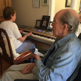 An elderly man watches as a woman plays the piano in a cozy indoor setting.
