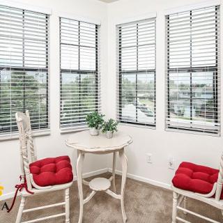 A cozy corner featuring a small table and chairs with red cushions, surrounded by large windows with blinds.