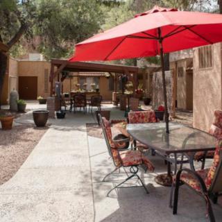 An outdoor patio area with a glass table, chairs, and a red umbrella set against a garden backdrop.