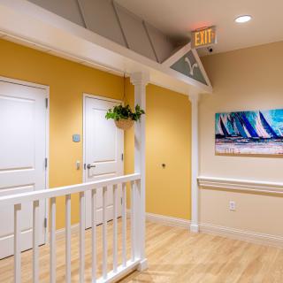 A bright hallway with yellow walls, white doors, and a wooden floor, featuring a painting and an exit sign.