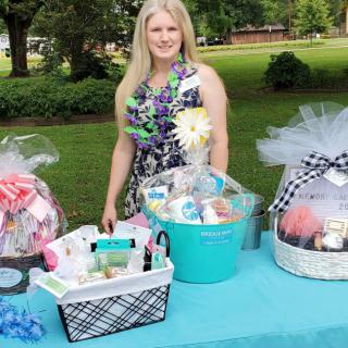 A woman stands behind a table with beautifully arranged gift baskets at an outdoor event.