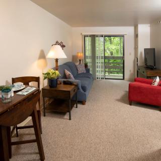 A cozy living room with a sofa, table, and television, featuring a view of greenery through sliding doors.