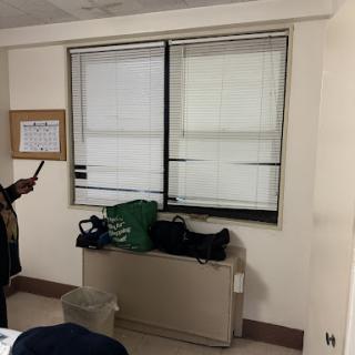 A person standing near the window in a room with blinds, a calendar on the wall, and some bags on a piece of furniture.