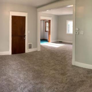 A spacious, empty living room with carpet flooring and light gray walls.