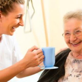 A caregiver assisting an elderly woman with a drink, both smiling happily.