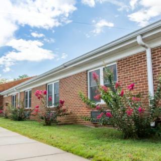 A red brick building with neatly maintained landscaping and blooming flowers, set against a partly cloudy sky.