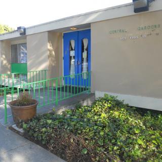 A building entrance with blue doors, surrounded by greenery and a plant pot.