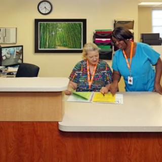 A caregiver assisting an elderly person at a nursing home reception desk.