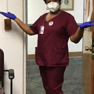 A healthcare worker in scrubs, wearing a face mask and gloves, greeting warmly.