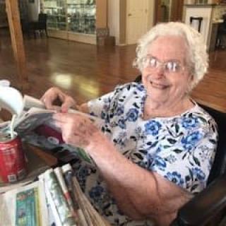 An elderly woman smiling while reading a newspaper indoors, with a soda can on the table.