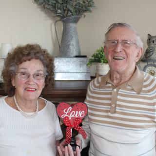 An elderly couple smiling and holding a red heart-shaped object with the word 'Love' on it.