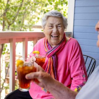 An elderly woman laughing while enjoying a drink on a balcony.