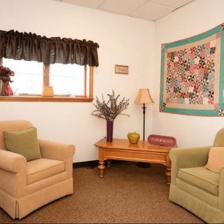 A cozy living room with two armchairs, a wooden table, and a wall-mounted quilt.