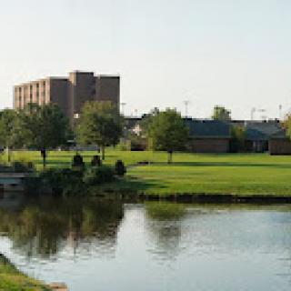 A serene view of a park area featuring greenery and a building in the background, with a calm body of water reflecting the scenery.