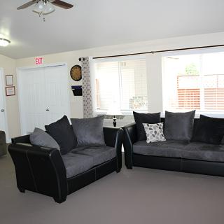 A bright living room featuring two dark sofas, a large window with curtains, and a ceiling fan.