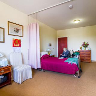 A hospital room with a single bed, where an elderly person is seated. The space includes a chair, dresser, and some wall art.