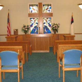 A small chapel with a stained glass cross, wooden pews, and flags on either side of the altar.