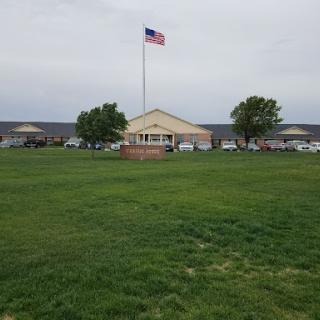 A view of a building with an American flag waving in front, surrounded by green grass and parked cars.