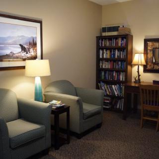 A cozy office space featuring comfortable chairs, a desk with a lamp, and a bookshelf filled with books.