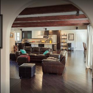A spacious living area with an open floor plan featuring a cozy seating arrangement, wooden beams, and a kitchen in the background.