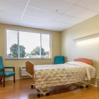A well-lit hospital room featuring a bed, two green chairs, and a window providing natural light.