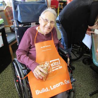 An elderly individual enjoying a community crafting activity while seated in a wheelchair, wearing an orange apron.